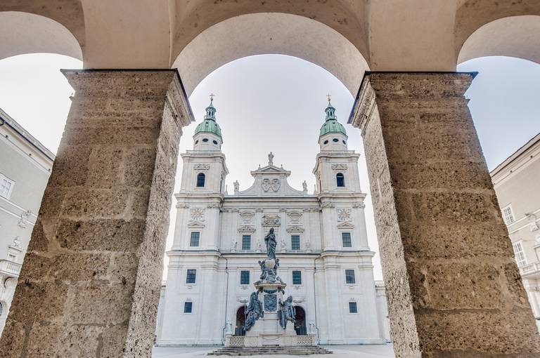 Blick auf eine historische Kathedrale mit zwei grün­kuppeligen Türmen, eingerahmt von steinernen Bögen, mit einer zentralen Statue im Vordergrund