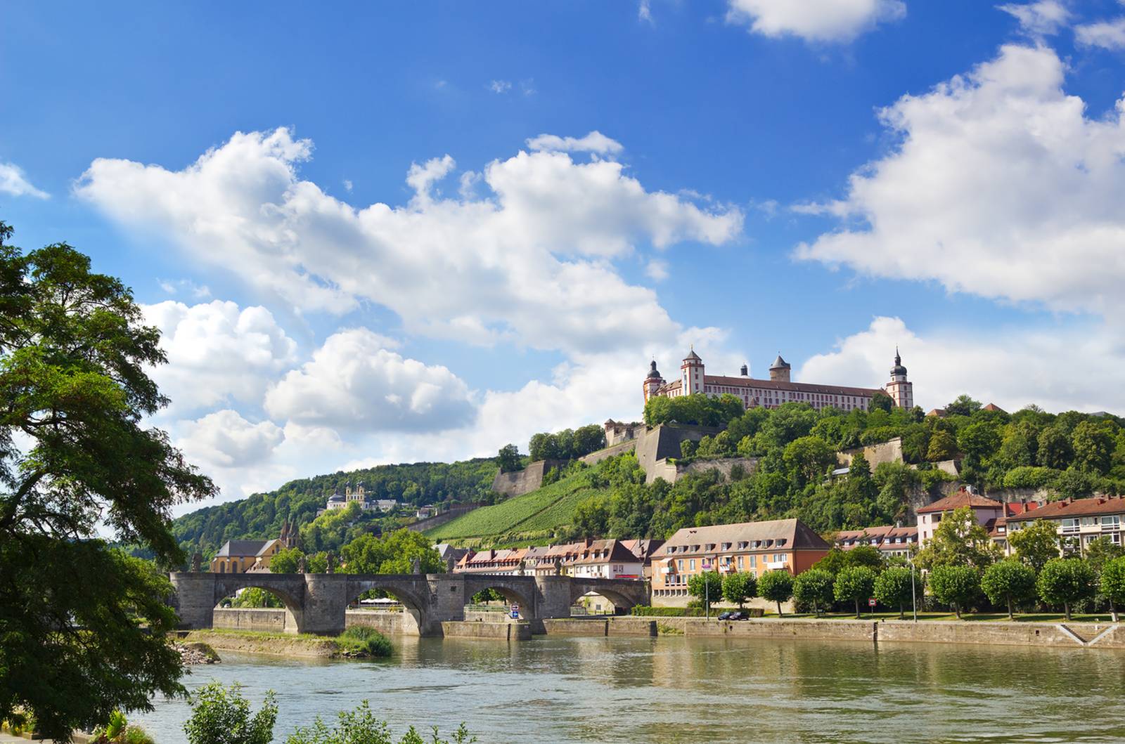 Malerischer Blick auf eine historische Festung auf einem Hügel, mit einer alten Steinbrücke, die einen Fluss überquert, und üppigem Grün unter blauem Himmel mit Wolken