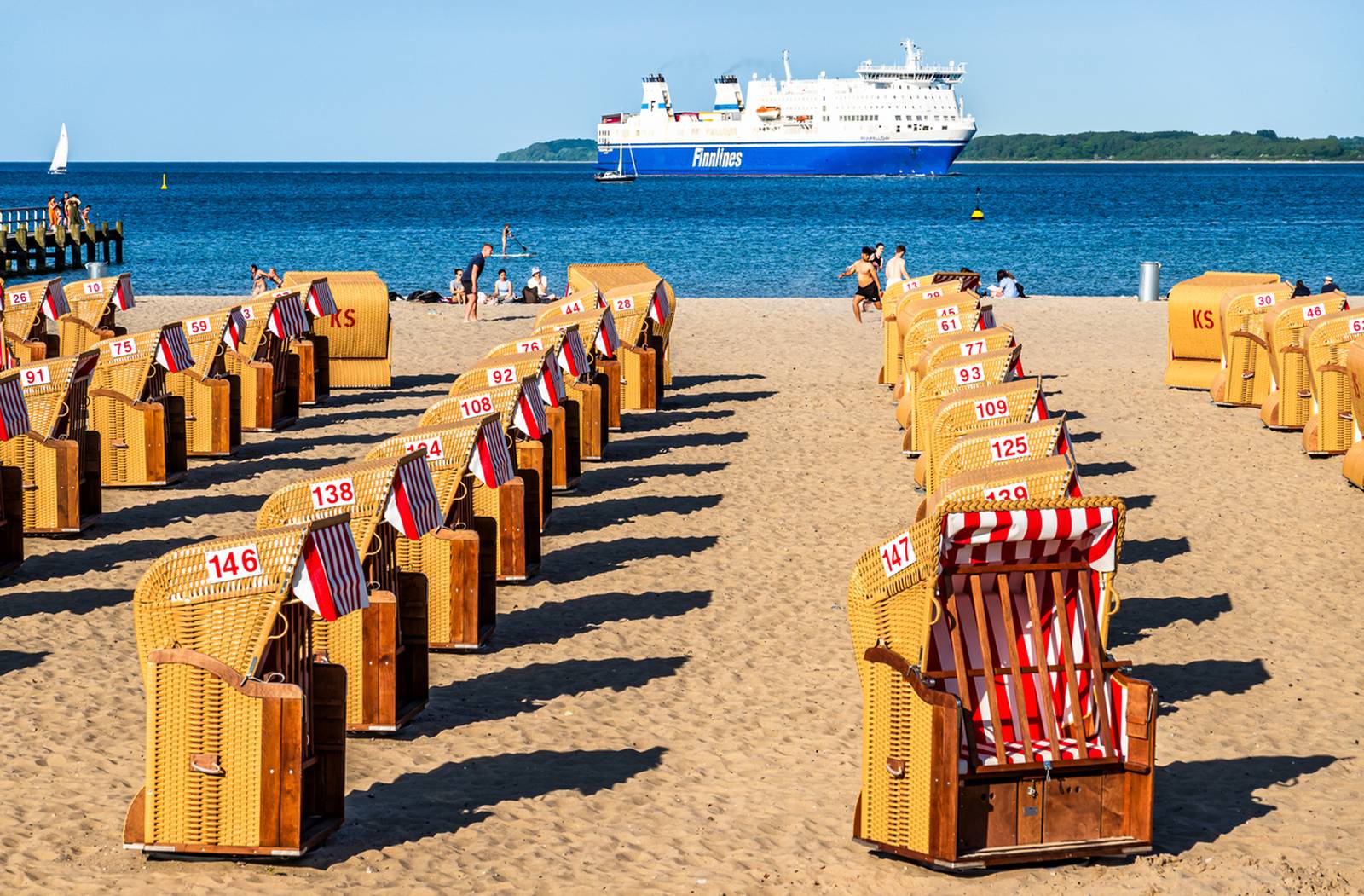 Strandszene mit Reihen von Strandkörben auf sandigem Untergrund, nummeriert und mit roten und weißen Streifen versehen. Im Hintergrund ist eine große Fähre auf dem blauen Meer zu sehen