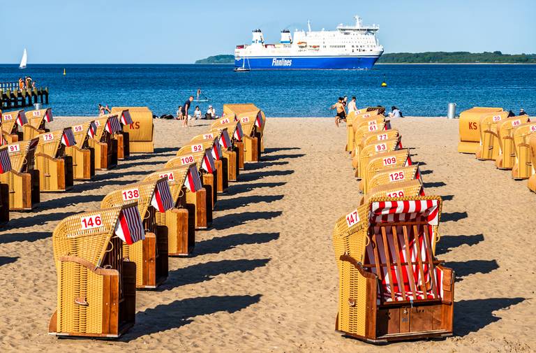 Strandszene mit Reihen von Strandkörben auf sandigem Untergrund, nummeriert und mit roten und weißen Streifen versehen. Im Hintergrund ist eine große Fähre auf dem blauen Meer zu sehen