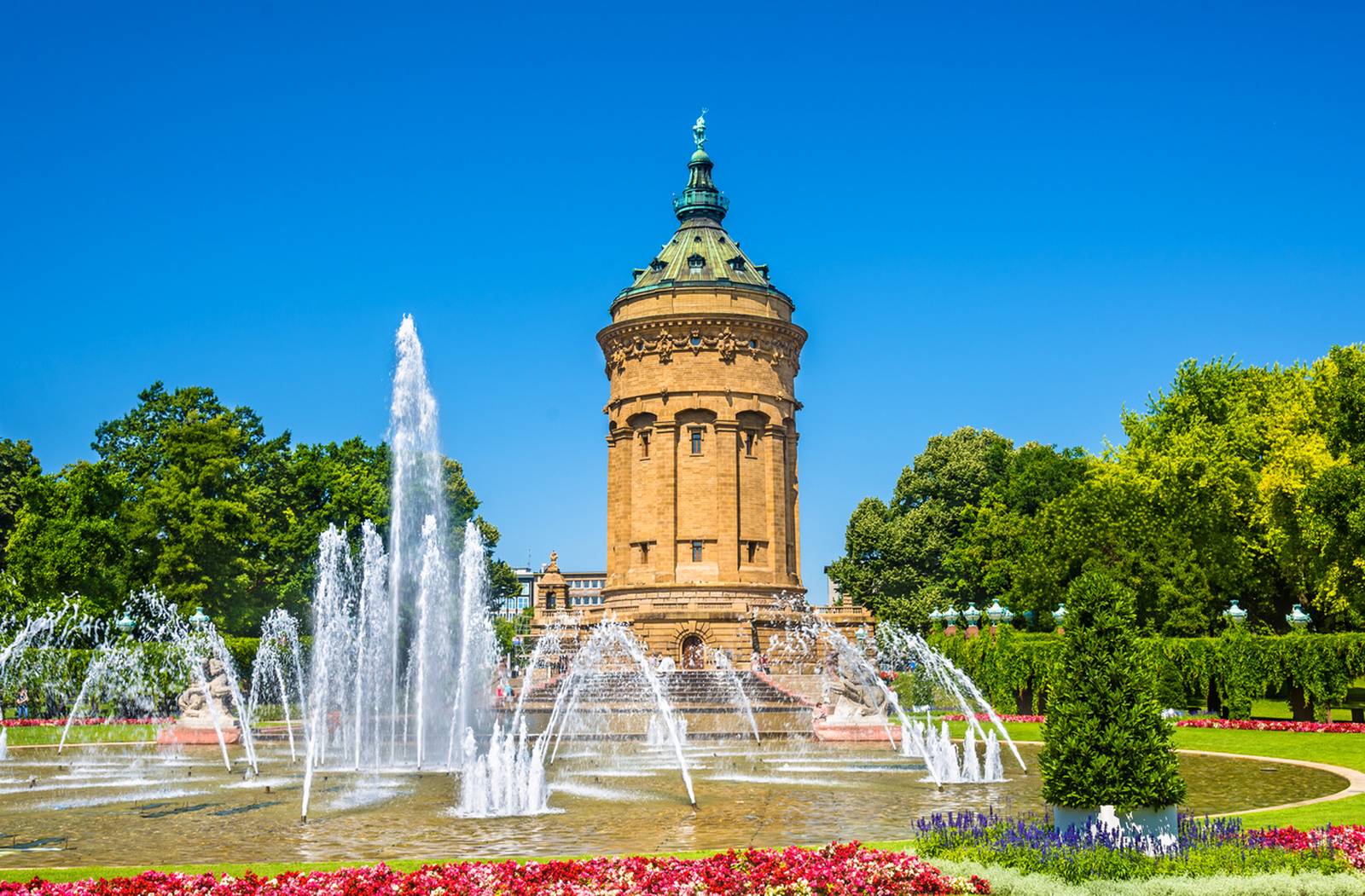 Historischer Wasserturm, umgeben von Springbrunnen und farbenfrohen Gärten unter klarem, blauem Himmel