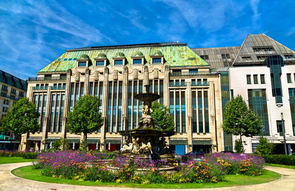 Prunkvoller Brunnen, umgeben von bunten Blumen, mit einem imposanten Gebäude im Hintergrund, das ein grünes Dach und gewölbte Fenster aufweist