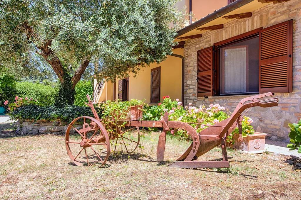 Agriturismo la morra assisi Old metal plow with wheels displayed in front of a stone house with wooden shutters, surrounded by greenery and a tree.