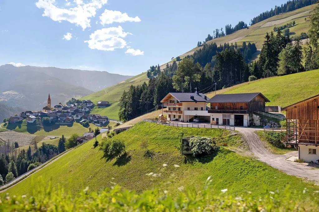 Agriturismo Alto Adige Mountain village with houses and a church on green slopes under a blue sky with clouds. Forests and fields surround the idyllic scene.