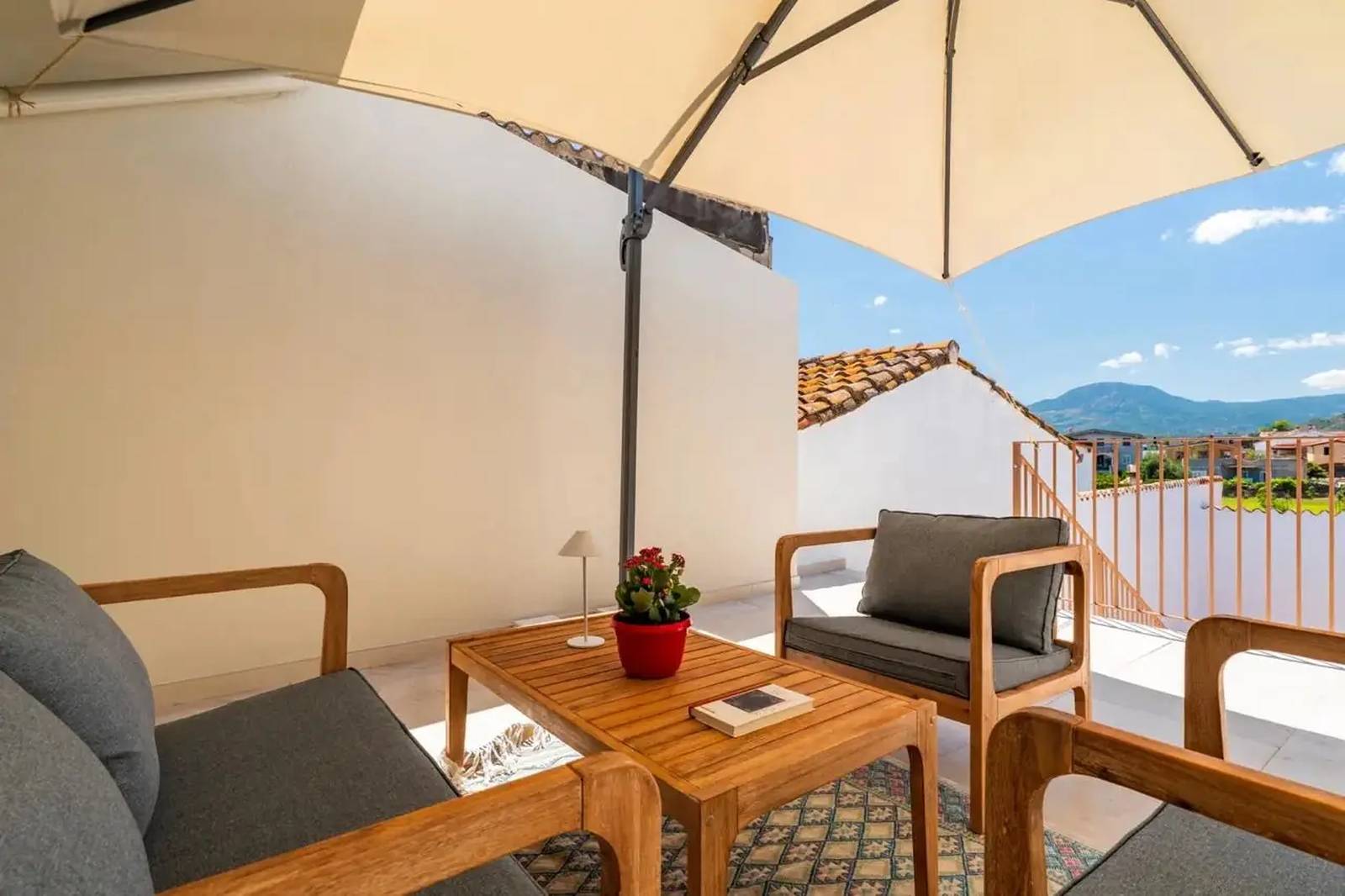 Dachterrasse mit zwei Holzstühlen, einem Tisch, einer Topfpflanze, einem Buch und Blick auf die Berge unter einem großen Sonnenschirm an einem sonnigen Tag.