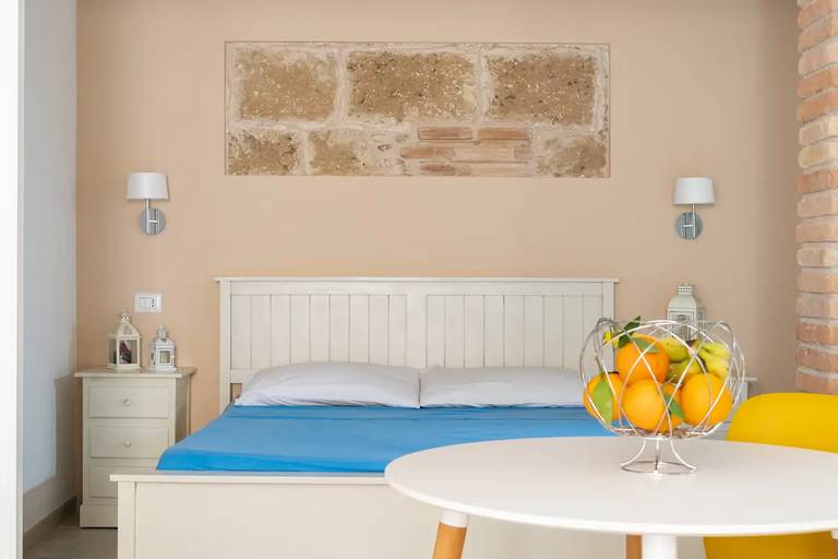 Gemütliches Schlafzimmer mit einem weißen Bett, blauer Bettwäsche, Nachttischen, einer Obstschale auf einem Tisch und Wandlampen vor einer strukturierten beige-farbenen Wand.