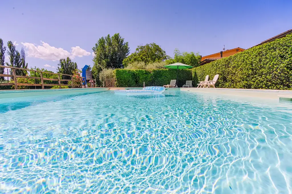 Clear pool with surrounding green hedge, lounge chairs, and a sun umbrella. Trees and a building with an orange roof in the background under a blue sky.