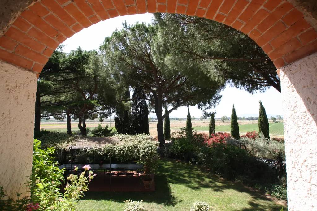 Brick archway framing a scenic view of lush trees, bushes, and a green lawn under a sunny sky, creating a tranquil garden setting.