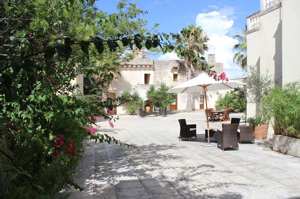 Masseria Pagani, Sunny courtyard with stone pavement, lush greenery, wicker chairs around a table under a white umbrella, and historic buildings in the background.
