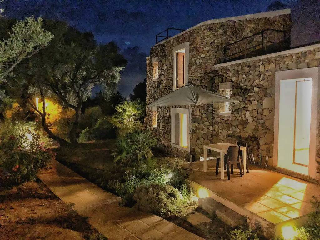 Ein Steinhaus mit beleuchteten Fenstern bei Nacht, mit einer Terrasse mit Sonnenschirm und Stühlen, umgeben von Bäumen und Gartenlichtern.