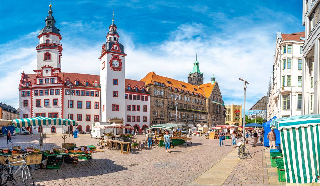 Sunny German town square with outdoor market stalls, bicycles and people in front of a historic red-roofed town hall and ornate buildings.