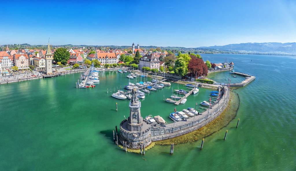 Luftaufnahme eines Yachthafens mit Booten, einem Leuchtturm, bunten Gebäuden und üppigem Grün unter klarem blauem Himmel.