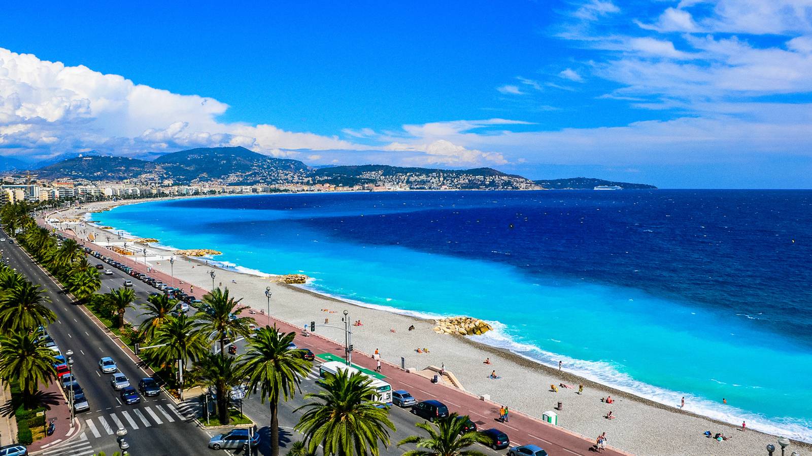 ChatGPT: Ein malerischer Blick auf eine Küstenstadt mit tiefblauem Meer, Sandstrand, palmengesäumter Promenade und fernen Bergen unter einem teilweise bewölkten Himmel