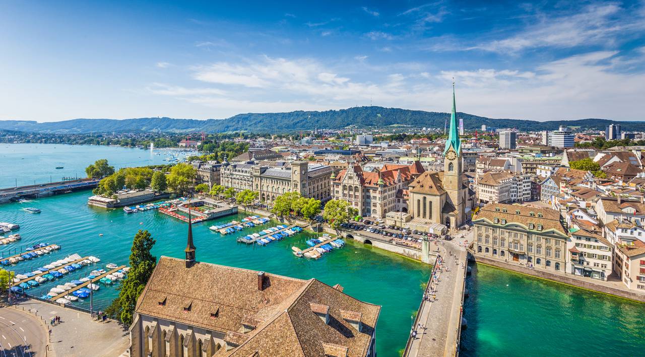 Luftaufnahme einer malerischen Stadtsilhouette mit einem Fluss, historischen Gebäuden, einer Kirche mit hohem Spitzturm, Booten und grünen Hügeln im Hintergrund.