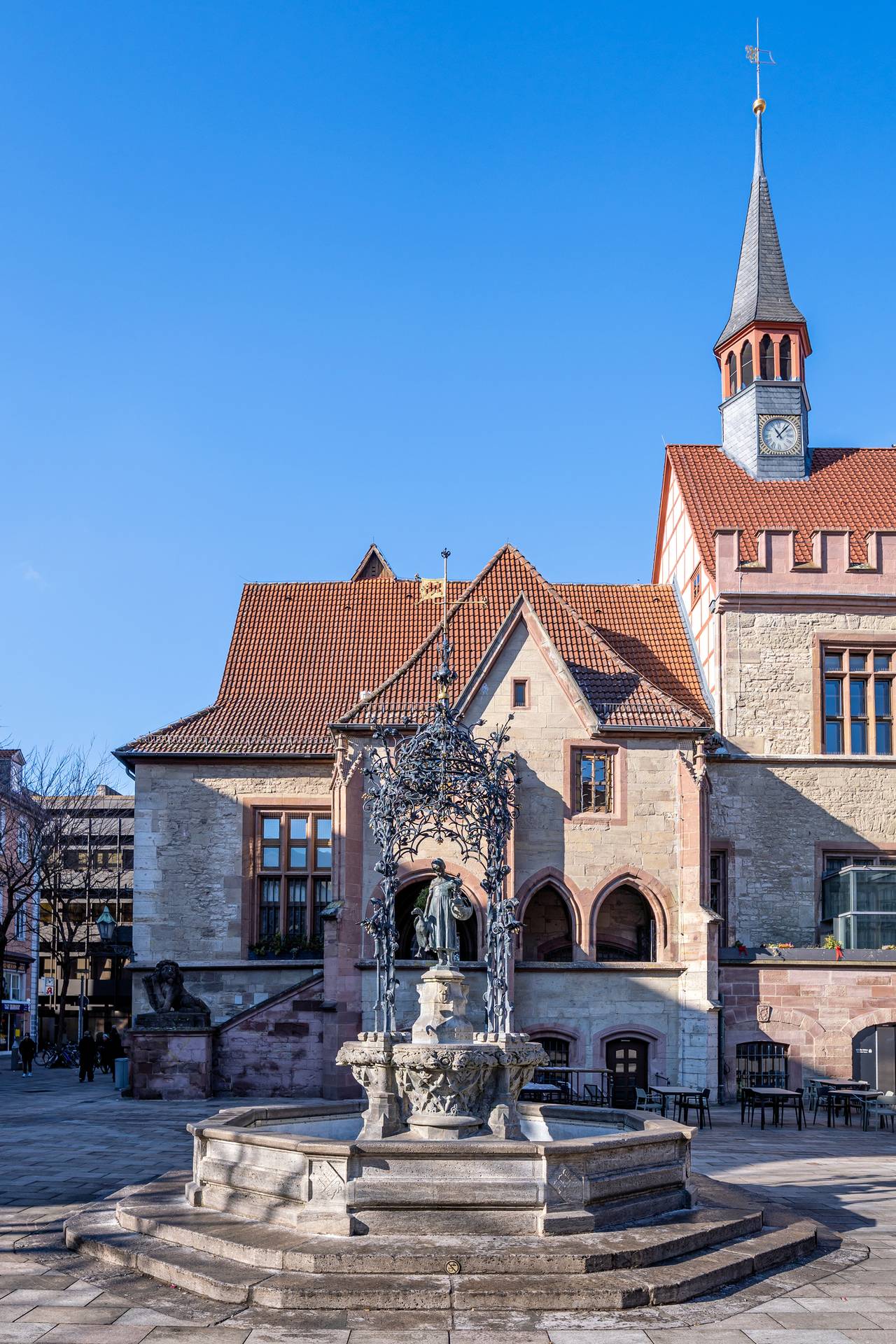 Historisches Gebäude mit Uhrturm und einem dekorativen Brunnen davor, vor klarem blauem Himmel