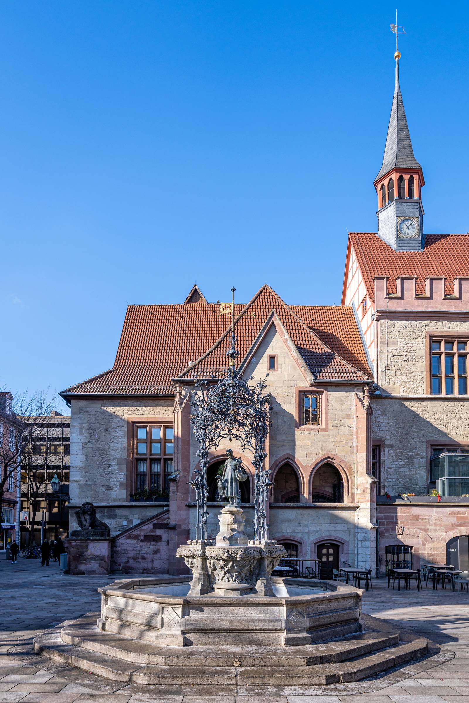 Historisches Gebäude mit Uhrturm und einem dekorativen Brunnen davor, vor klarem blauem Himmel