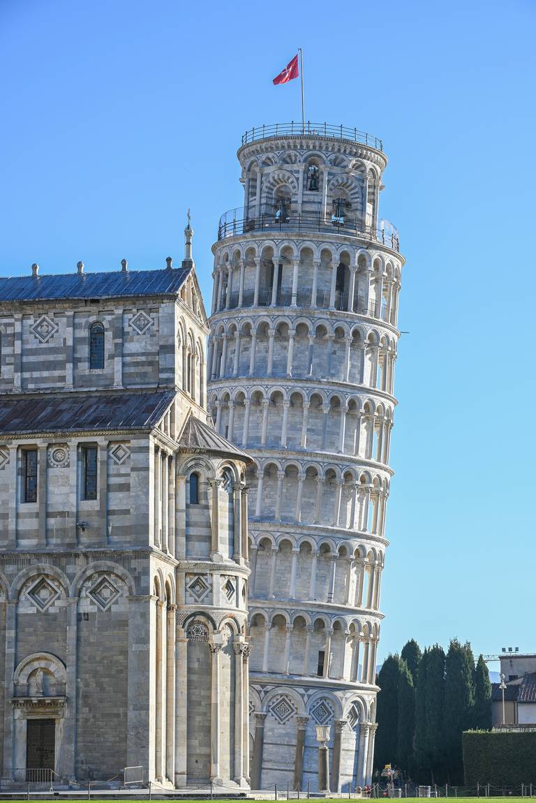 Der schiefe Turm von Pisa mit einer roten Flagge auf der Spitze, neben einem historischen Gebäude, vor einem klaren blauen Himmel.