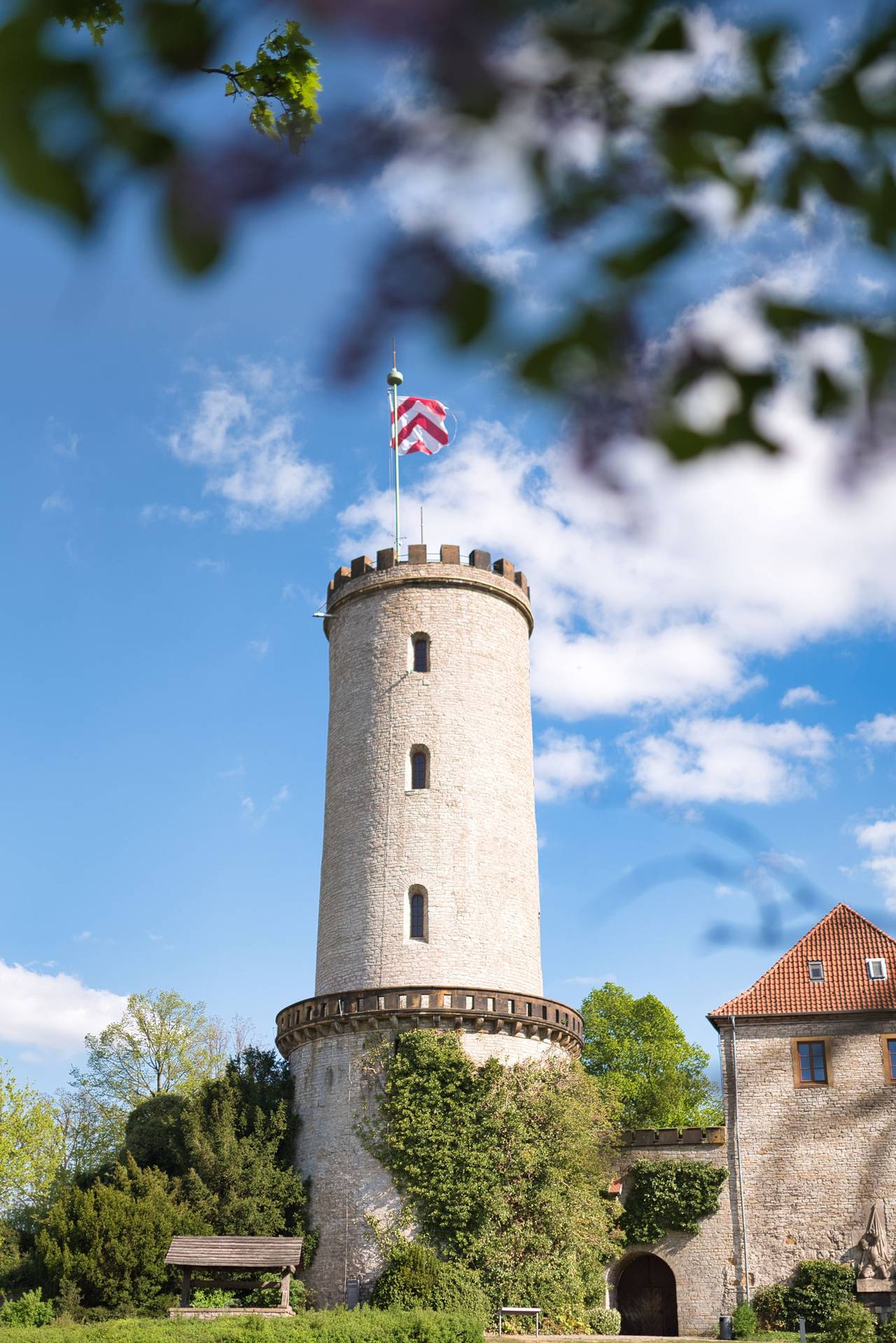 Hoher Steinturm mit Zinnenkranz und rot-weißer Fahne, umgeben von grüner Vegetation und blauem Himmel mit vereinzelten Wolken