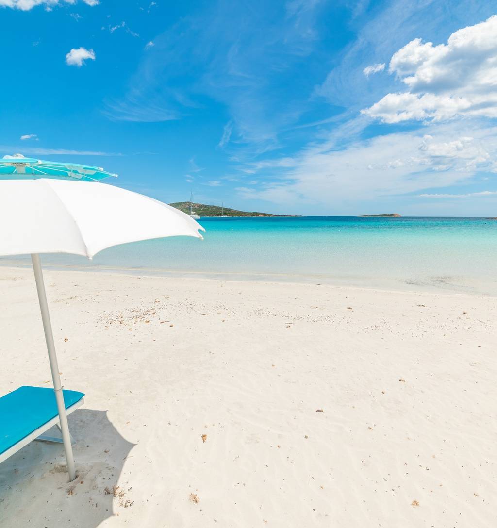 Weißer Sandstrand mit türkisfarbenem Meer unter blauem Himmel. Ein weißer Sonnenschirm spendet Schatten für eine blaue Liege. Am Horizont ist fernes Land zu sehen.