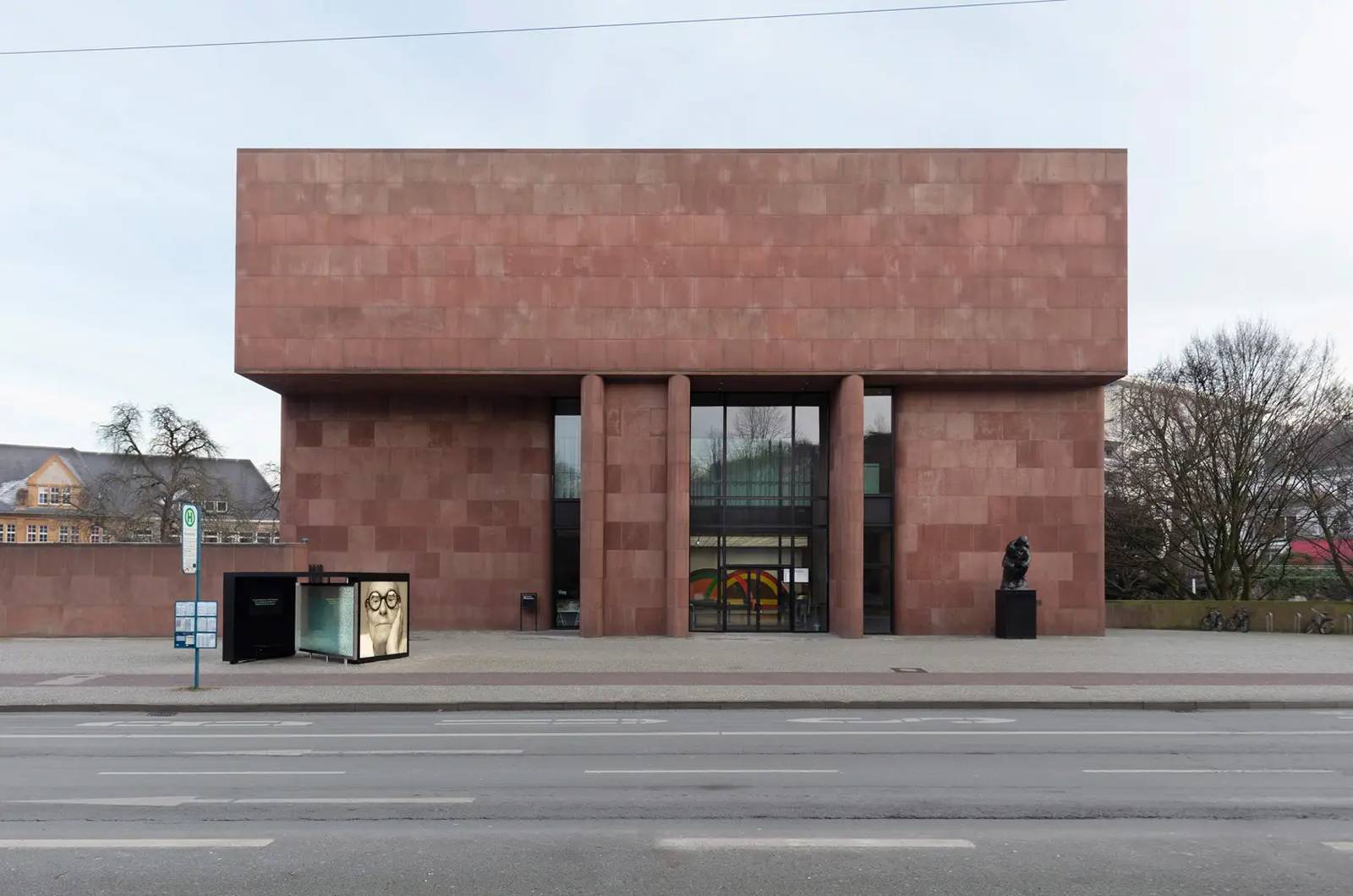 Modernes Gebäude mit roter Ziegelfassade, großem gläsernen Eingangsbereich und zwei senkrechten Säulen, gelegen an einer Straße mit nahegelegenem Busbahnhof