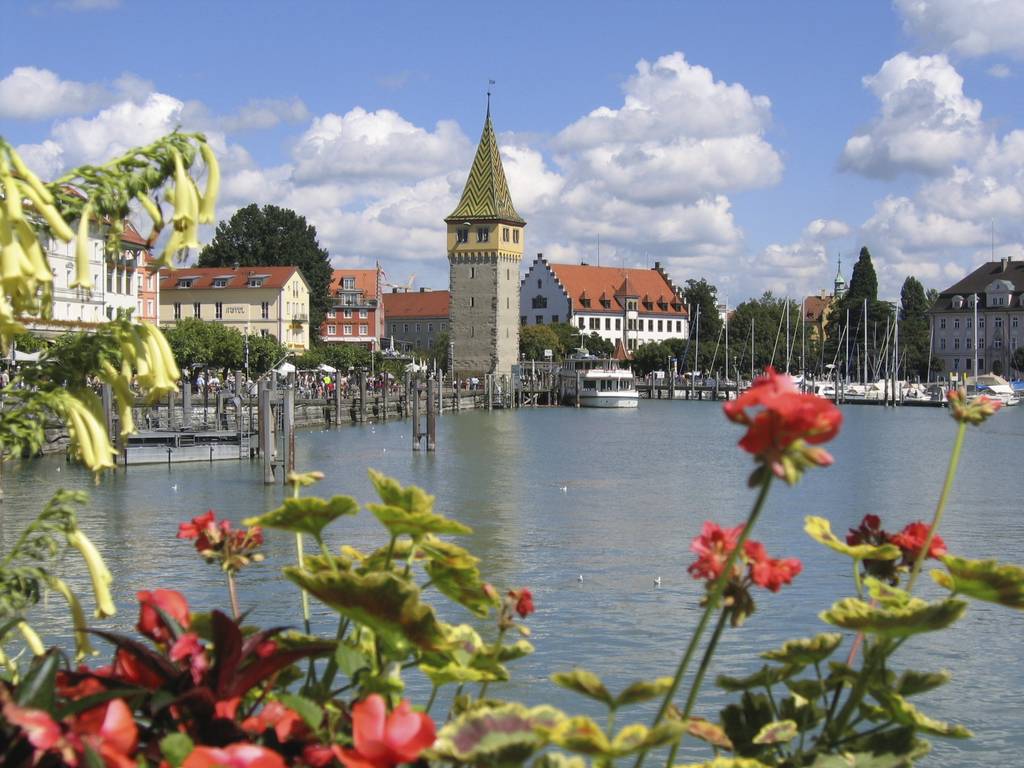 Malerischer Bodensee mit historischem Turm, bunten Blumen im Vordergrund und Booten am Steg, unter einem blauen Himmel mit Wolken.