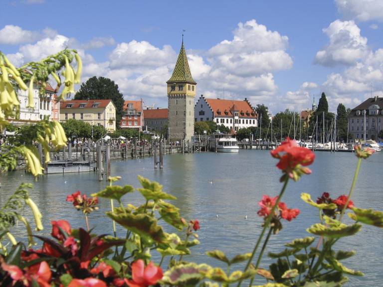 Städtchen am See mit einem steinernen Turm und einem Ziegeldachspitze, Segelboote im Hafen, rote Blumen im Vordergrund und blauer Himmel mit fluffigen Wolken.