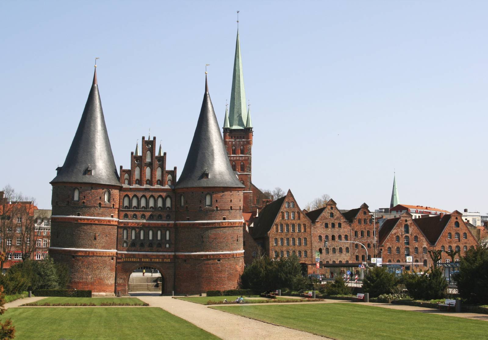 Ein historisches Backsteintor mit zwei konischen Türmen steht vor klarem, blauem Himmel, umgeben von grünen Rasenflächen und alten Gebäuden