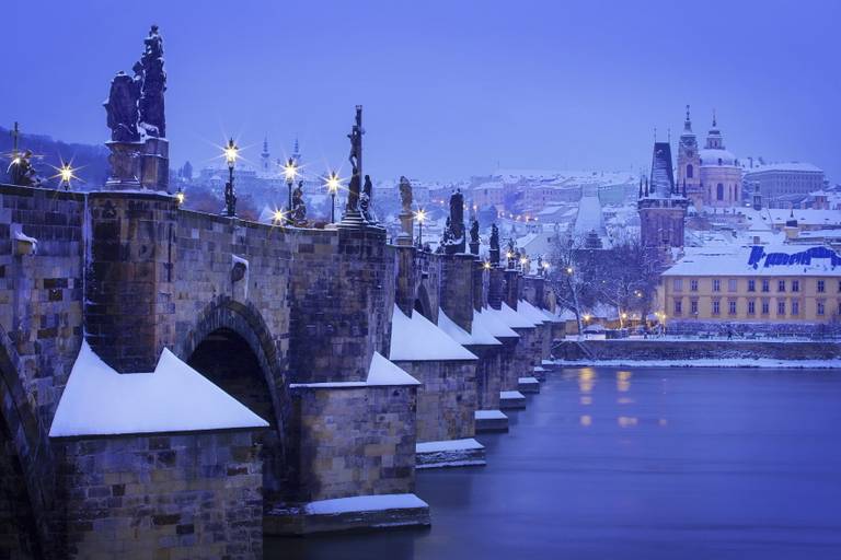Die verschneite Karlsbrücke in Prag in der Dämmerung, mit beleuchteten Statuen und historischen Gebäuden im Hintergrund, deren Spiegelung sich in der Moldau zeigt.