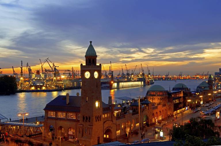 Sonnenuntergang über einem belebten Hafen: beleuchteter Uhrenturm am Fluss, Hafenkräne als Silhouette vor einem farbenfrohen Himmel und spiegelnde Lichter.