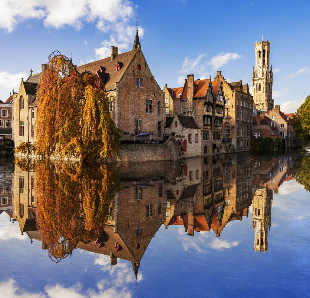 Historic buildings with gabled roofs reflected in a calm canal, framed by an autumn tree and a tower under a blue sky with clouds.