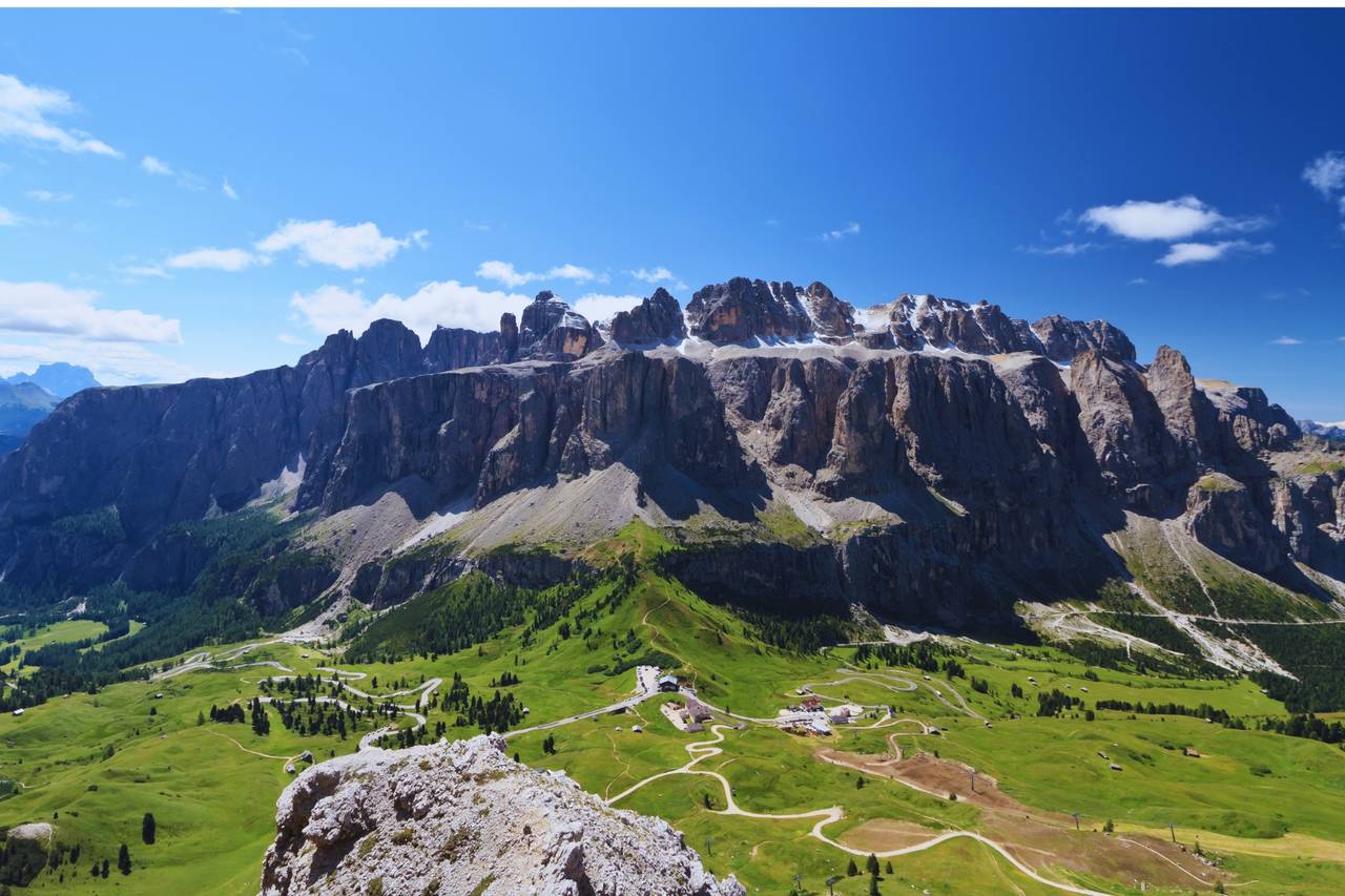 Majestätische Bergkette unter einem strahlend blauen Himmel mit vereinzelten Wolken, umgeben von üppig grünen Wiesen und sich schlängelnden Straßen im Tal darunter.