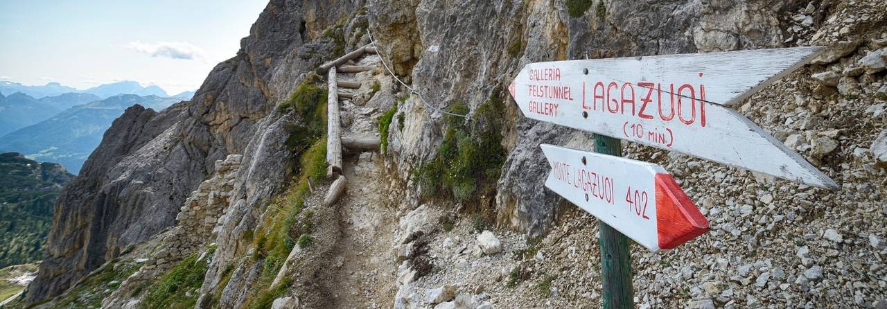 Bergpfad mit einer hölzernen Leiter, die bergauf führt. Wegweiser zeigen in Richtung Lagazuoi und geben Wanderziele sowie Entfernungen an. Felsiges Gelände mit Blick auf die Berglandschaft.