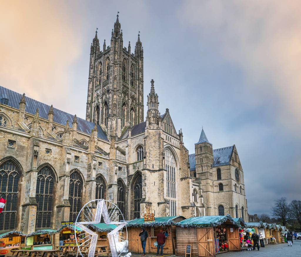 Canterbury Cathedral with a cloudy sky, surrounded by festive market stalls displaying colorful goods.