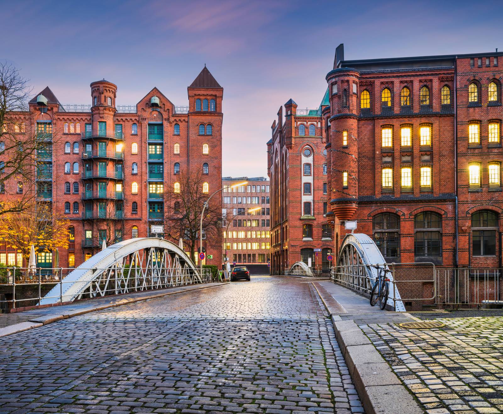 Kopfsteinpflasterstraße mit einer Brücke, die zu historischen Backsteingebäuden mit beleuchteten Fenstern führt, unter einem rosafarbenen Himmel in der Dämmerung