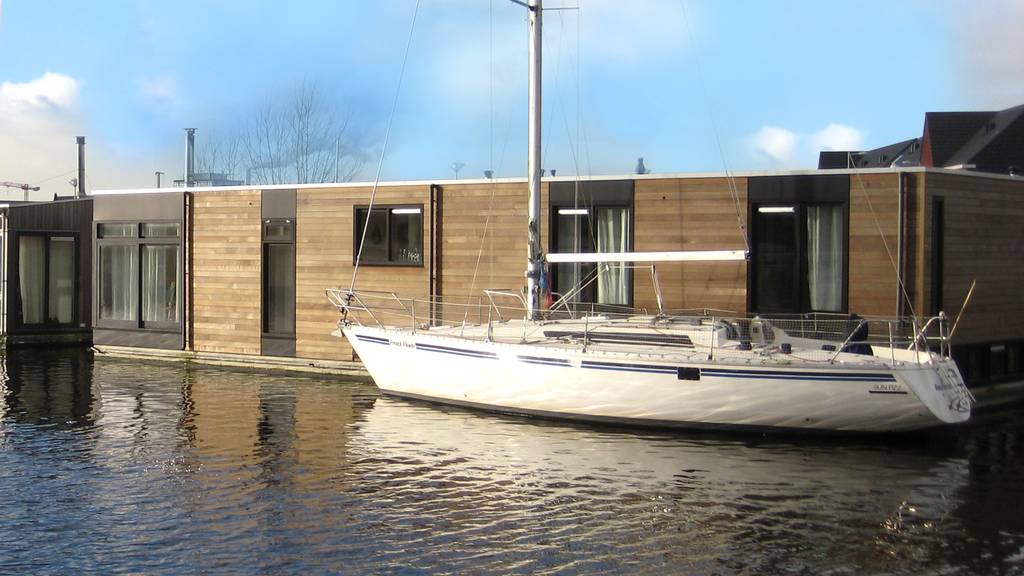 Modern houseboat with wooden exterior and large windows, moored alongside a canal, with a sailboat docked nearby under a clear blue sky.