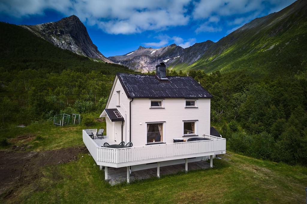 Une maison blanche dotée d'une terrasse qui fait le tour de la maison, nichée dans une vallée verdoyante entourée de montagnes, sous un ciel bleu partiellement nuageux.