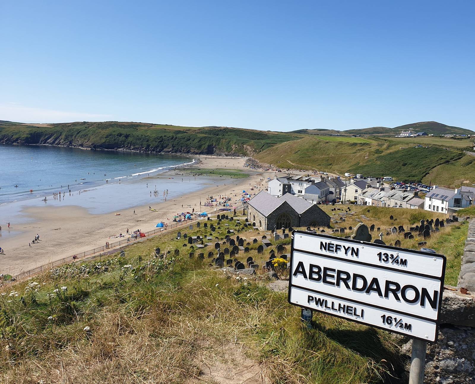 Coastal village view with sandy beach, people sunbathing, lush green hills, and a signpost for Aberdaron, Nefyn, and Pwllheli.