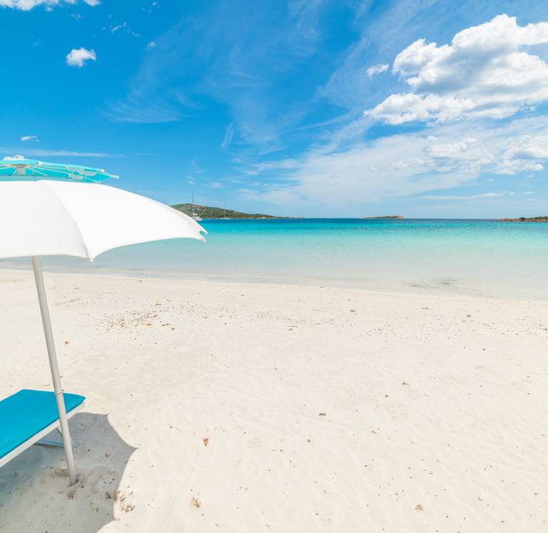 Sunny beach of Sardinia scene with a white umbrella and blue lounge chair on the sand, overlooking turquoise ocean water and a clear blue sky.