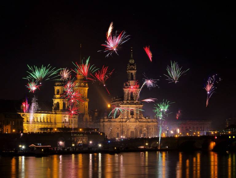Feuerwerk erleuchtet den Nachthimmel über einem beleuchteten historischen Gebäude am Fluss, die bunten Farben spiegeln sich lebendig im Wasser.