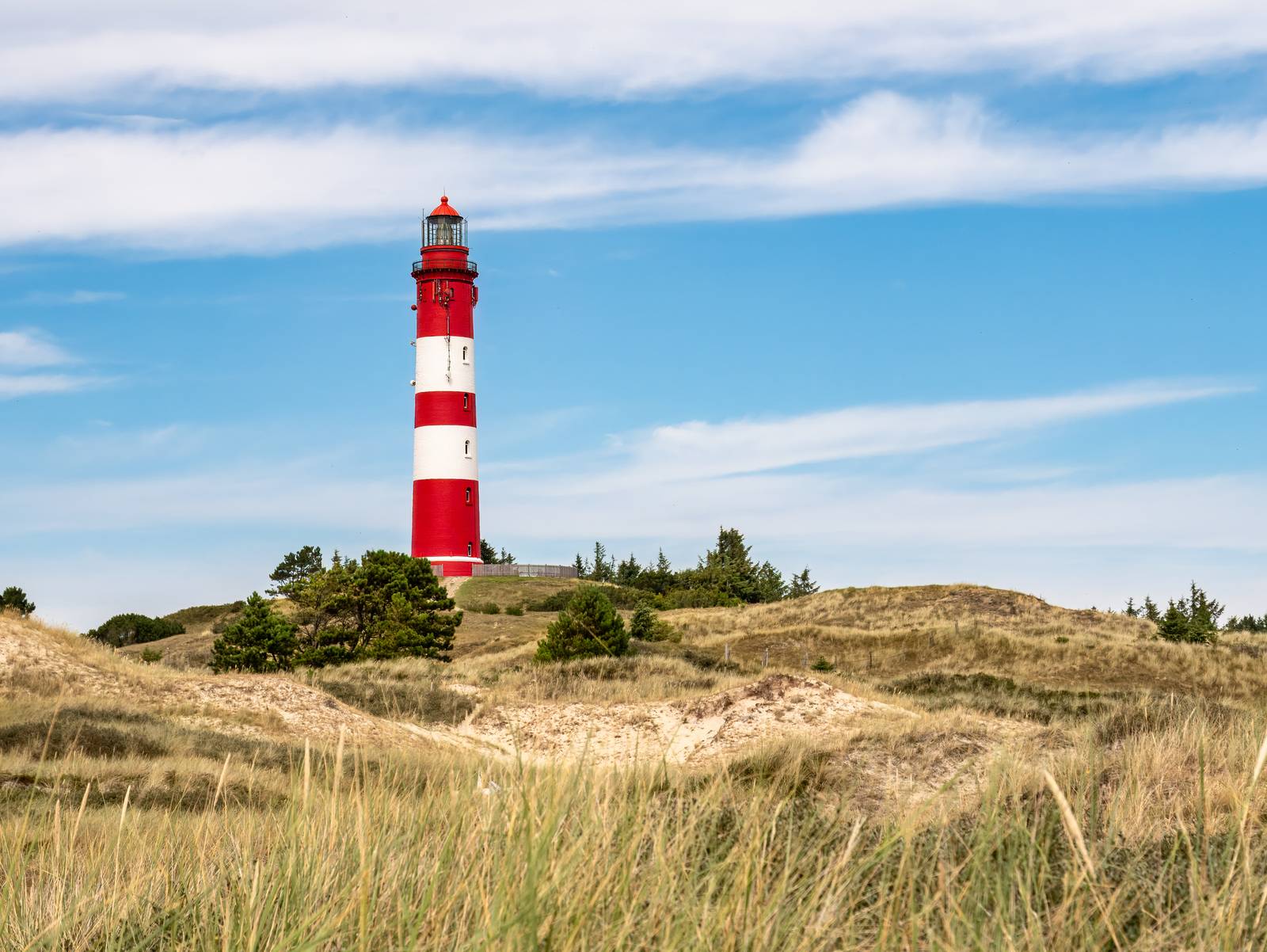 Rot-weiß gestreifter Leuchtturm auf einer grasbewachsenen Düne unter einem blauen Himmel mit vereinzelten Wolken.