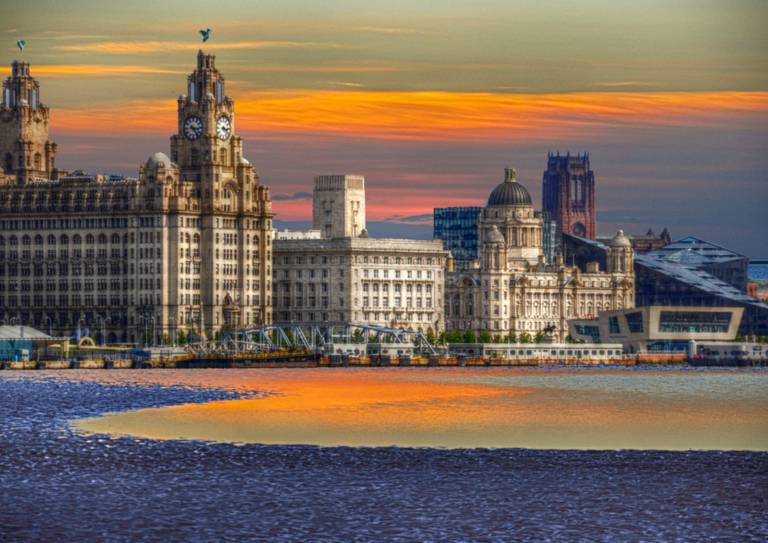 Three Graces: Skyline of Liverpool featuring historic buildings at sunset, with vibrant orange and yellow hues reflected on the water.
