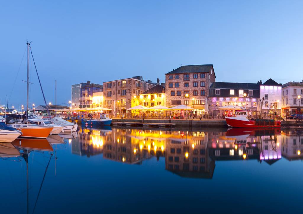 Plymouth old port at twilight with boats, lit buildings reflecting on calm water, and clear blue sky in the background.