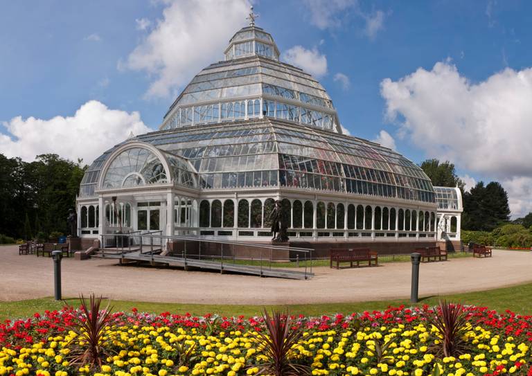 Sefton Park Palm House, A large glass conservatory with a dome roof, surrounded by colorful flowerbeds, under a partly cloudy sky.