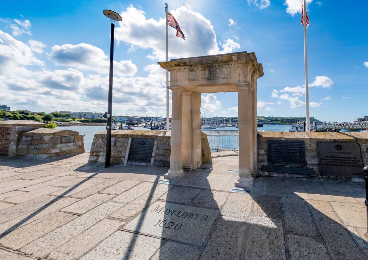 mayflower steps plymouth Stone arch overlooking a marina, flanked by Union Jack flags and plaques. The ground bears the inscription "Mayflower 1620." Bright, sunny day.