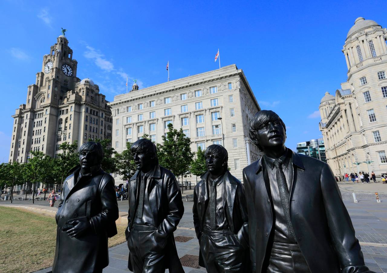 Statues of the beatles in liverpool, with historic buildings and a clock tower in the background under a clear blue sky.