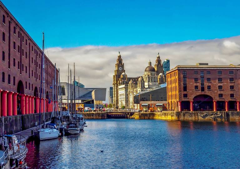 Historic waterfront with red brick buildings in Liverpool, moored boats, and iconic clock towers under a bright blue sky.