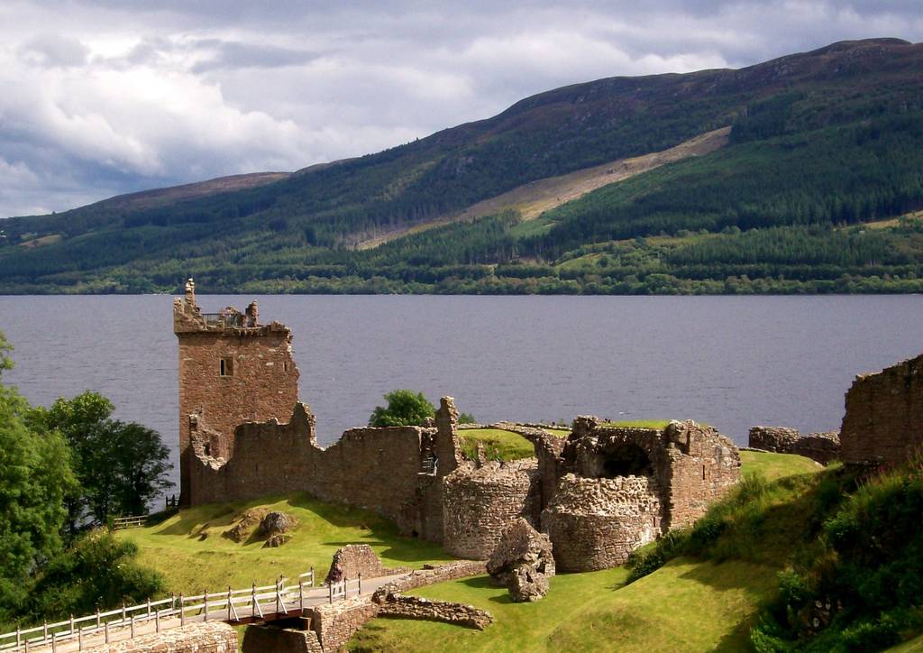 Ruins of  Urquhart Castle by the shore of a large loch, surrounded by lush green hills under a cloudy sky.