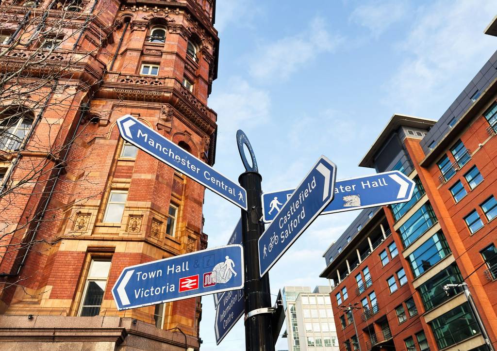 Street signs in a city center, directing to Manchester Central, Castlefield, Albert Hall, and Town Hall, with historic and modern buildings behind.