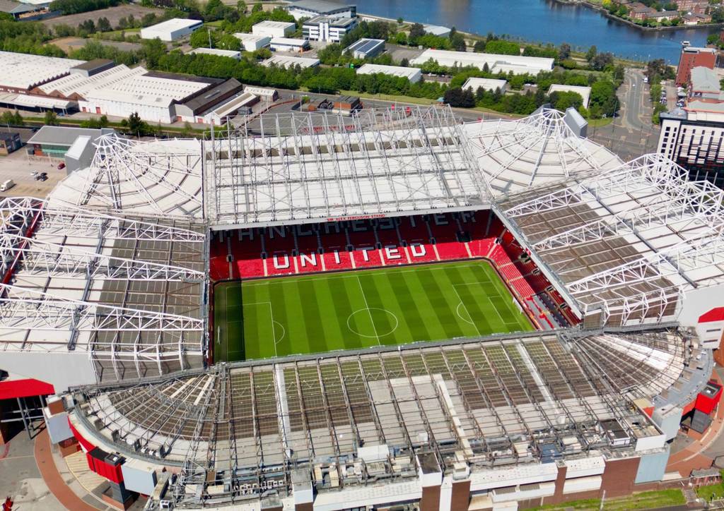 Aerial view of a large football stadium Old Trafford, Manchester,  with a green pitch, surrounded by seating. The roof has a lattice design, and buildings are visible in the background.