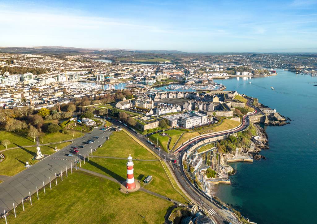 Aerial view of plymouth with a prominent lighthouse, grassy parkland, winding coastal road, and expansive waterfront under a clear blue sky.